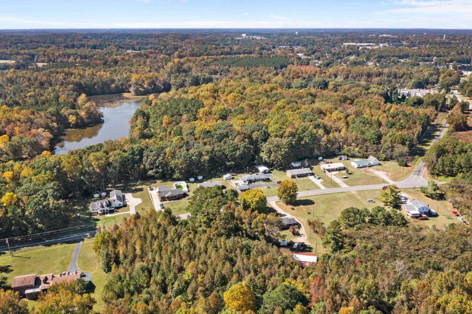 Aerial view of a suburban area with houses and roads amidst a landscape with a mix of evergreen and deciduous trees showing autumn colors, with a river meandering in the background.