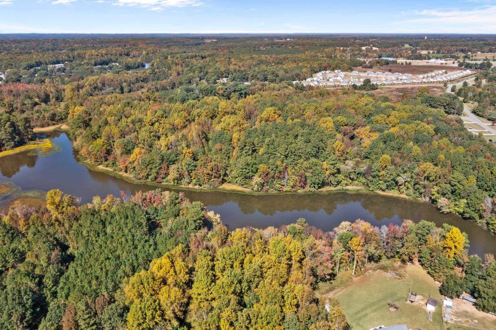 Aerial view of a meandering river surrounded by trees with autumn foliage, with a clear sky above and developed land near the horizon.