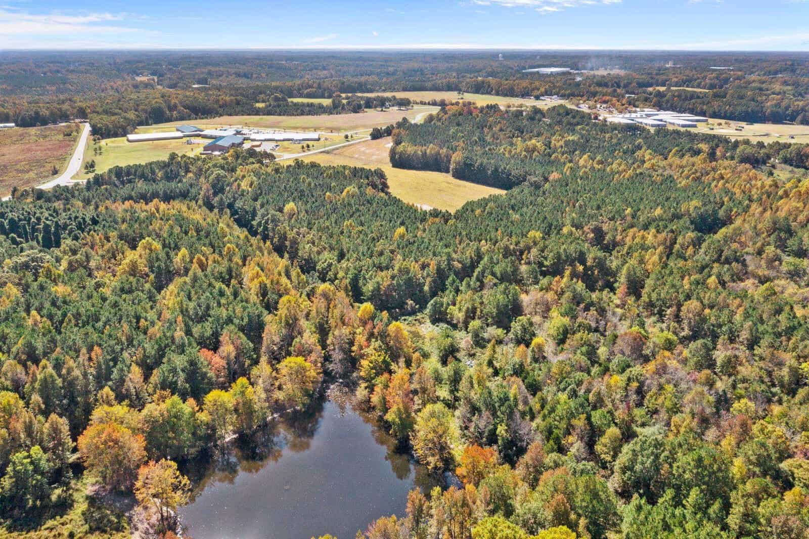 Aerial view of a landscape showing a mix of autumn-colored woodland, a small lake, open fields, and distant buildings under a clear sky.