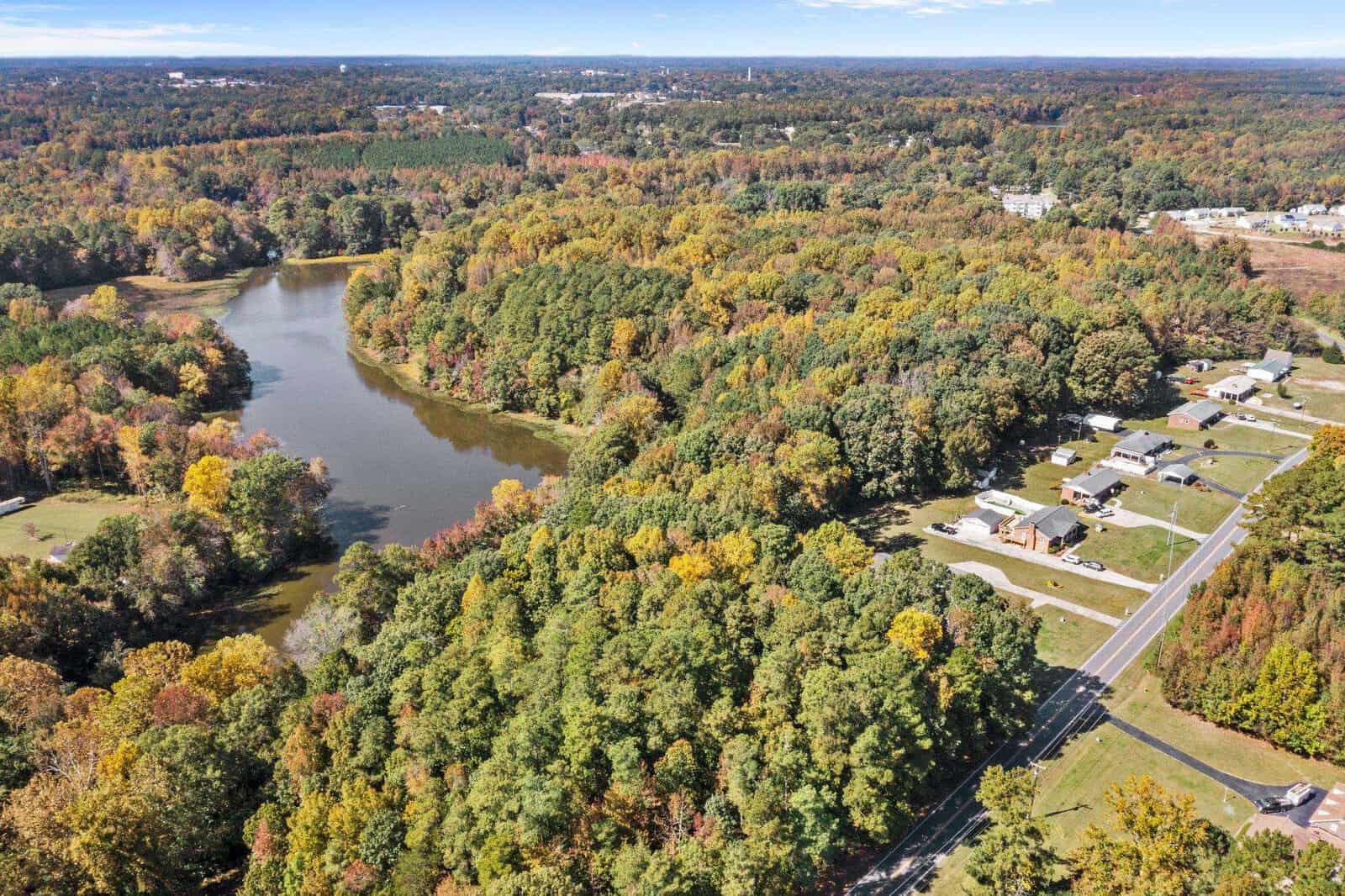 Aerial view of a suburban area with houses alongside a road, surrounded by a landscape of trees showcasing autumn foliage and a small lake nearby.