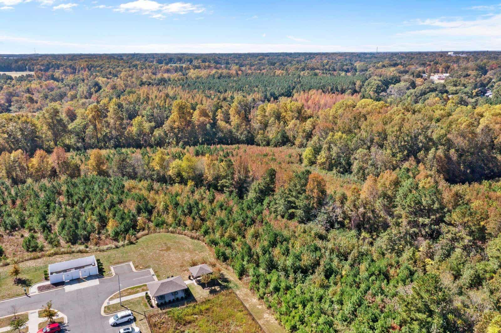 Aerial view of a rural landscape showing a mix of deciduous trees in autumn colors and evergreens, with a few buildings and a parking area surrounded by nature.