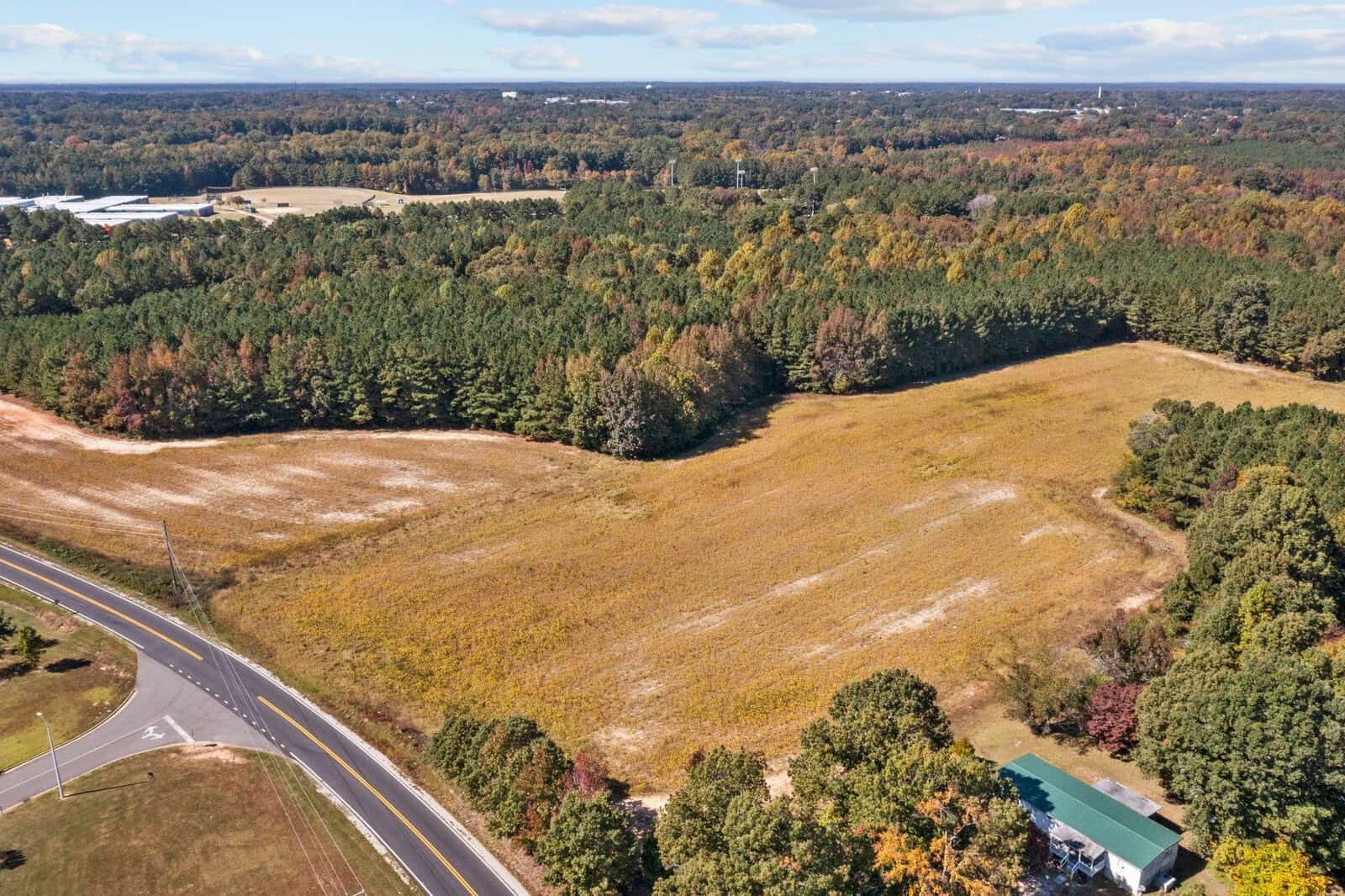 Aerial view of a cleared field adjacent to a forest with a road running parallel to it and a small building on the bottom right corner.