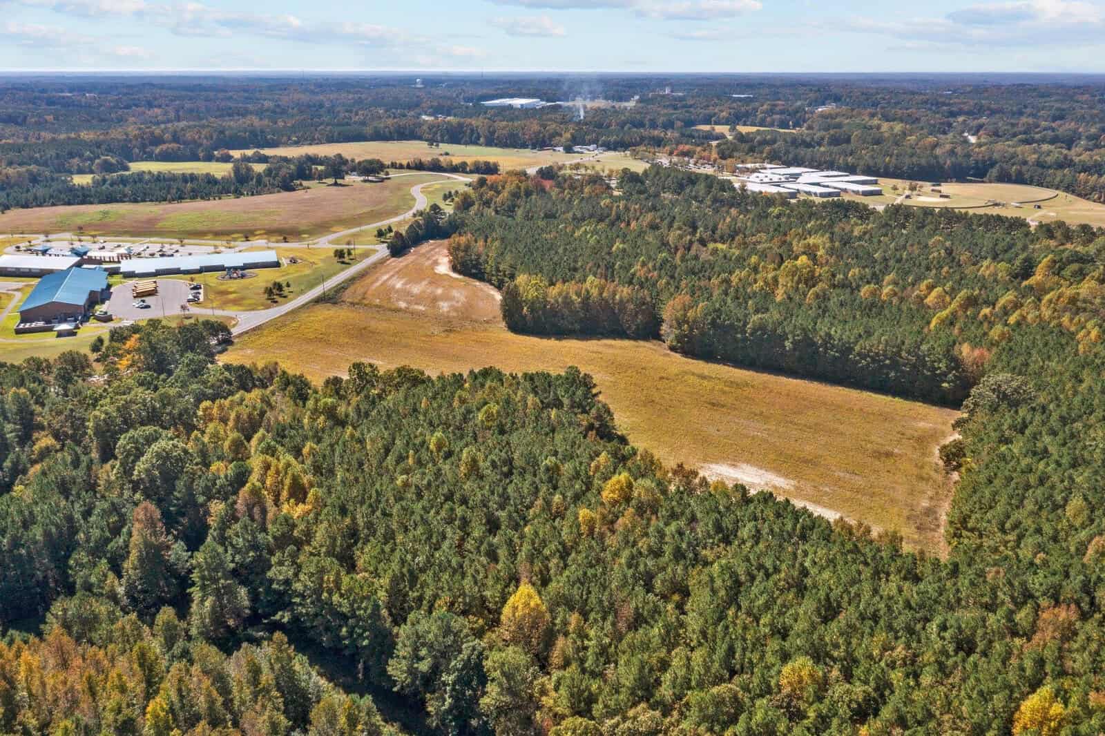 Aerial view of a rural landscape showing a mix of wooded areas, open fields, and industrial buildings along a winding road.