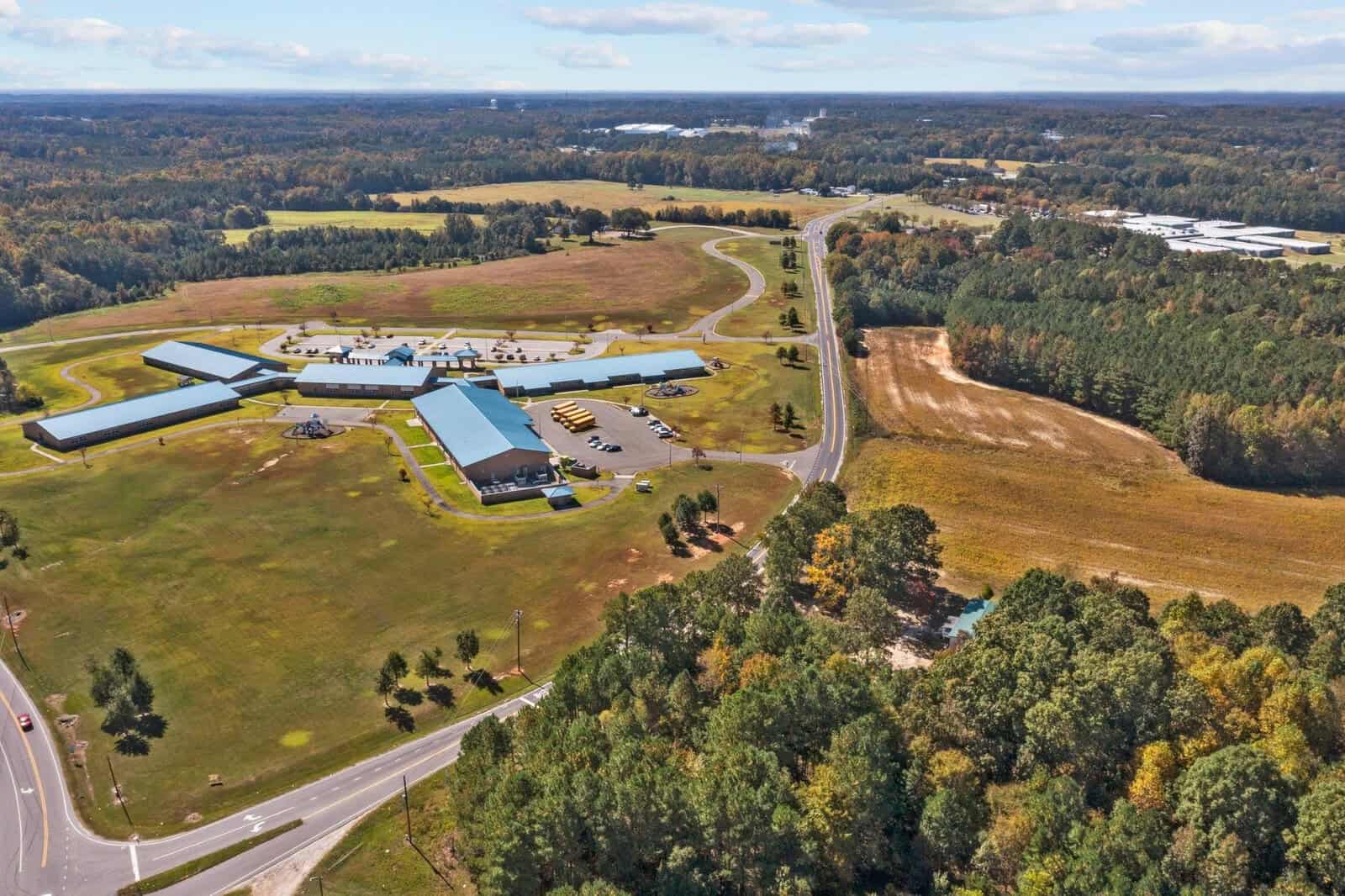 Aerial view of a rural landscape featuring a large building complex with a blue roof, surrounded by expansive green fields, roads, and patches of forest, under a partly cloudy sky.