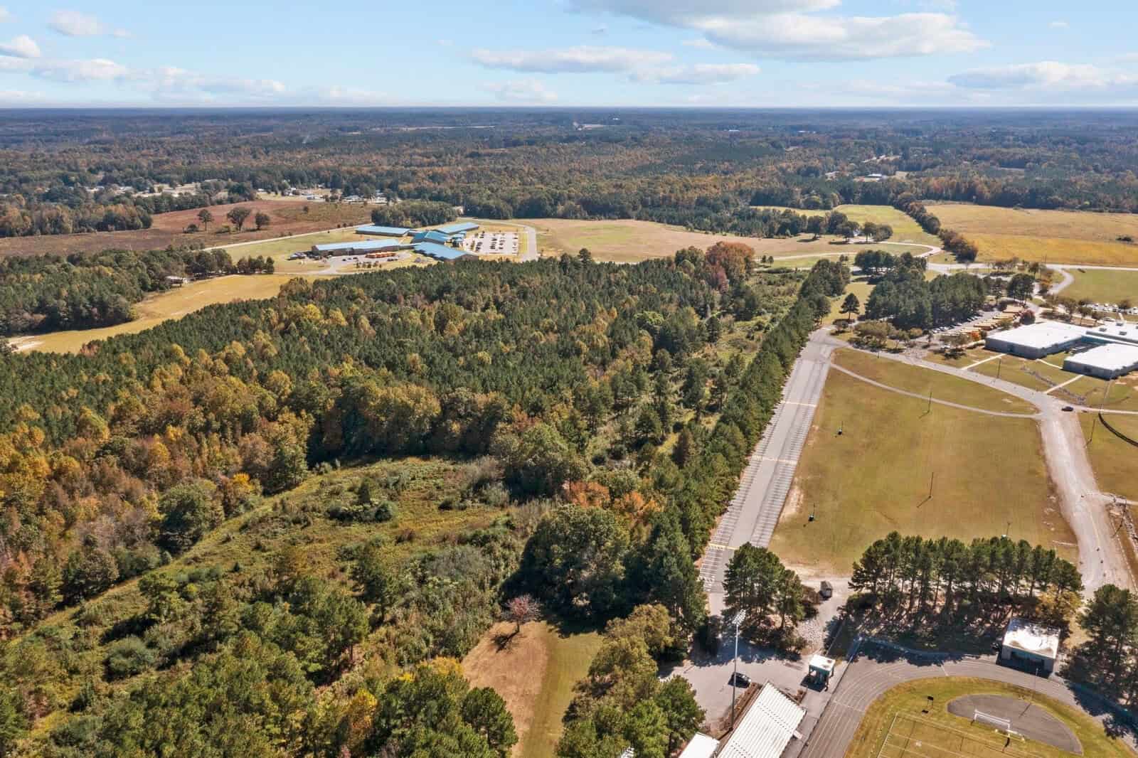 Aerial view of a rural landscape featuring patches of forest, fields, and scattered buildings, with a train track running parallel to a road.
