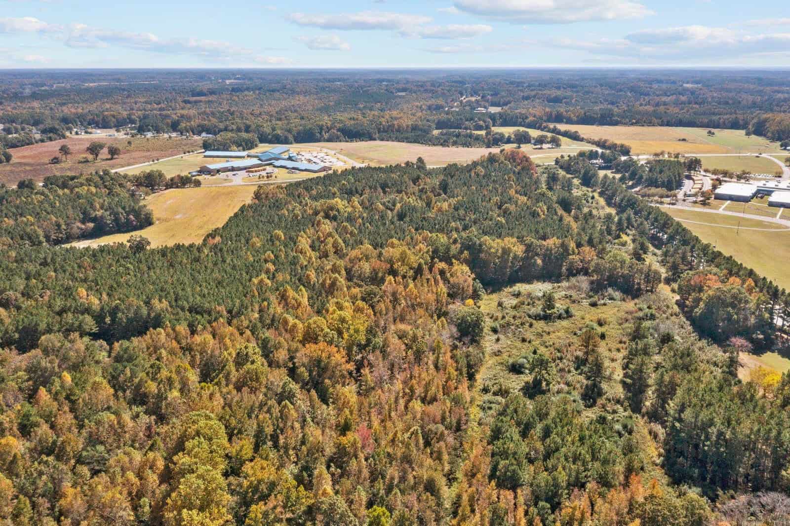 Aerial view of a varied landscape showing patches of forest with autumnal foliage adjacent to open fields, with buildings in the distance under a partially cloudy sky.