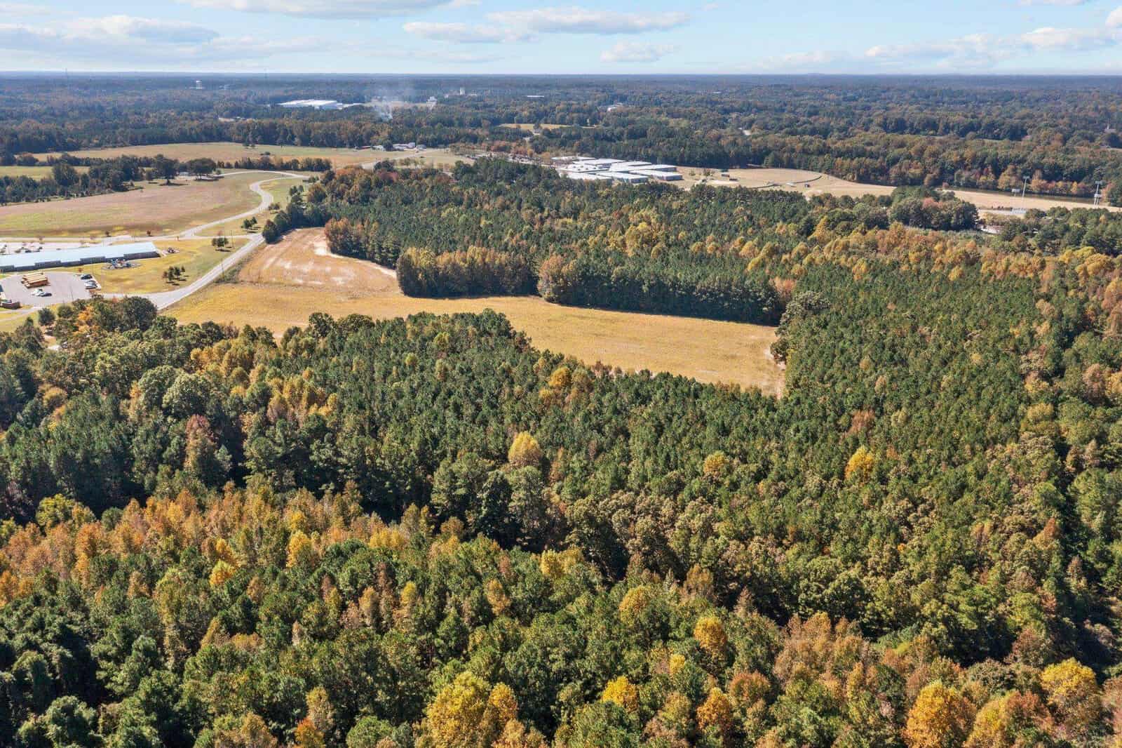 Aerial view of a landscape showing a mix of forested areas with fall foliage and open land, with a road and buildings in the distance.