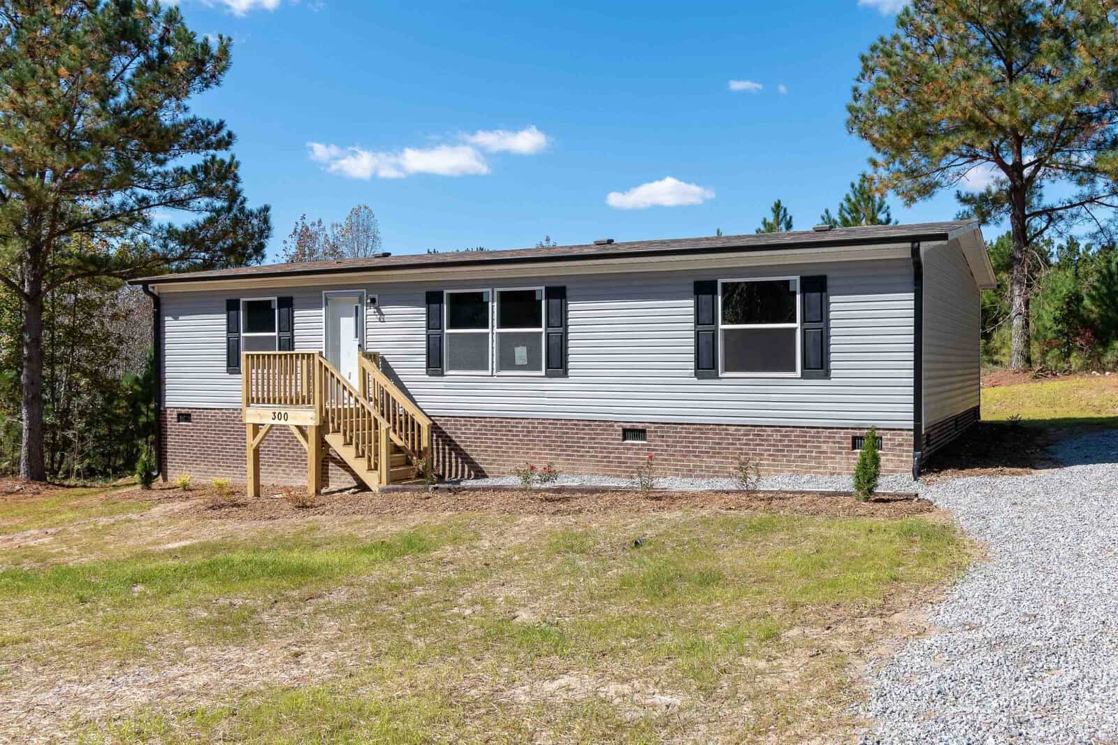 A single-story manufactured home with gray siding, white trim, and a small wooden porch, located in a clearing with a background of trees.