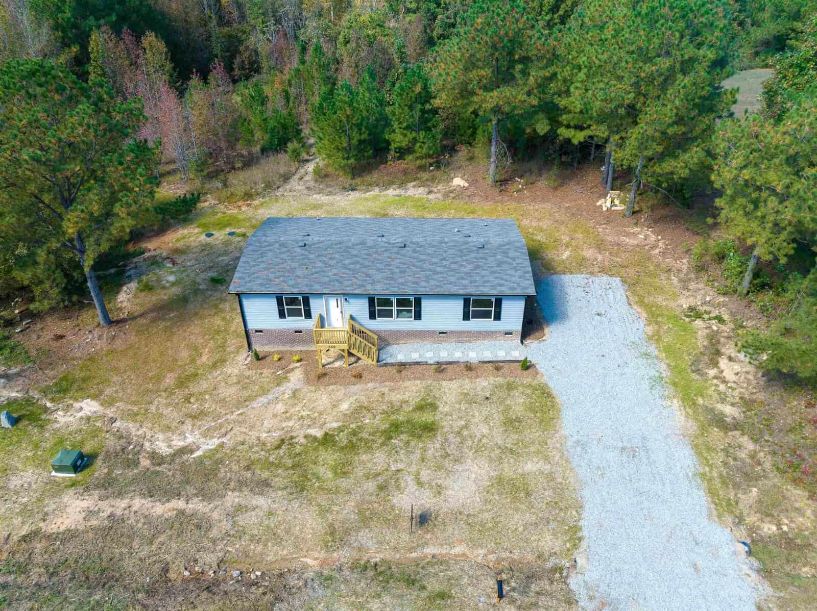Aerial view of a single-story house with a gray roof, surrounded by sparsely wooded land and a gravel driveway.