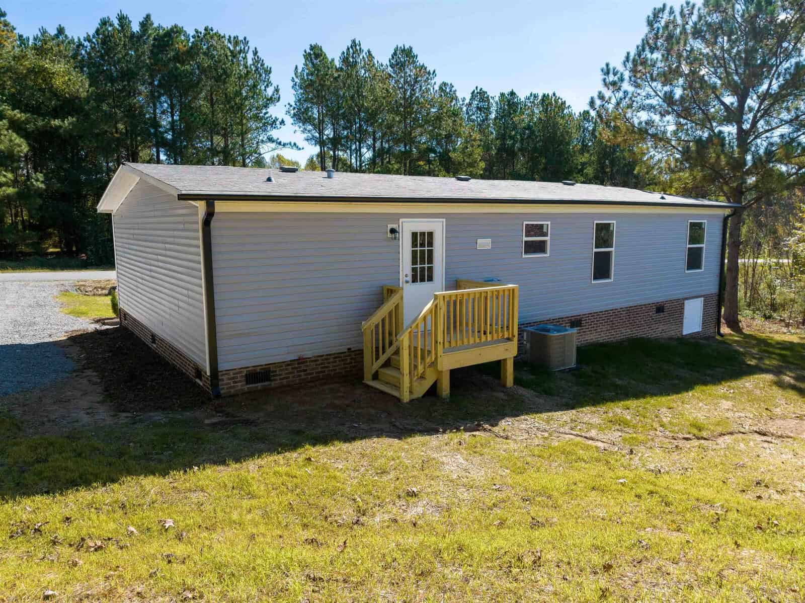 A single-story modular home with vinyl siding, featuring a small wooden deck with steps, situated in a grassy area with surrounding trees.