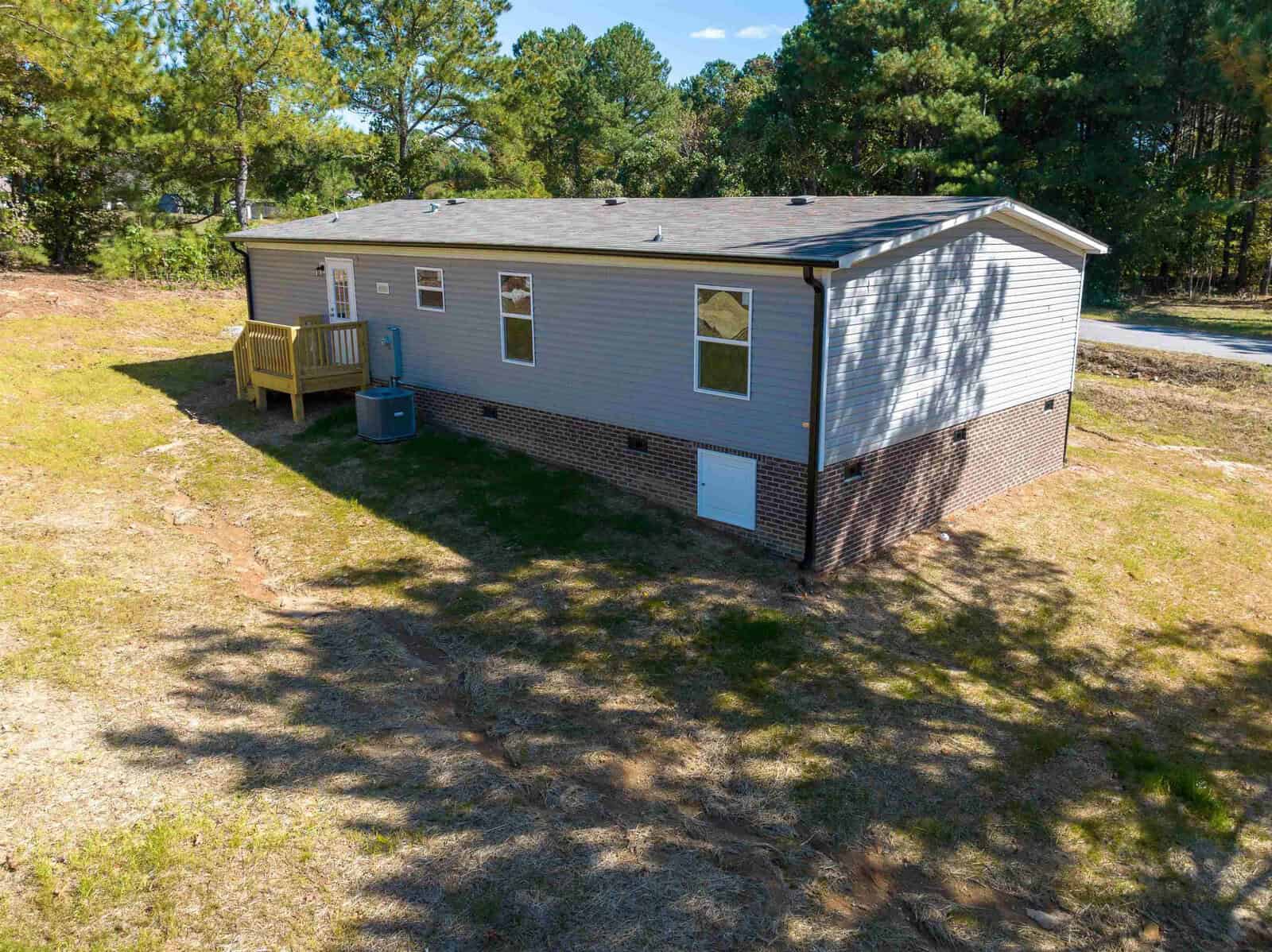Aerial view of a single-story modular home with gray siding and a small wooden deck, situated on a cleared lot with sparse grass and surrounding trees.