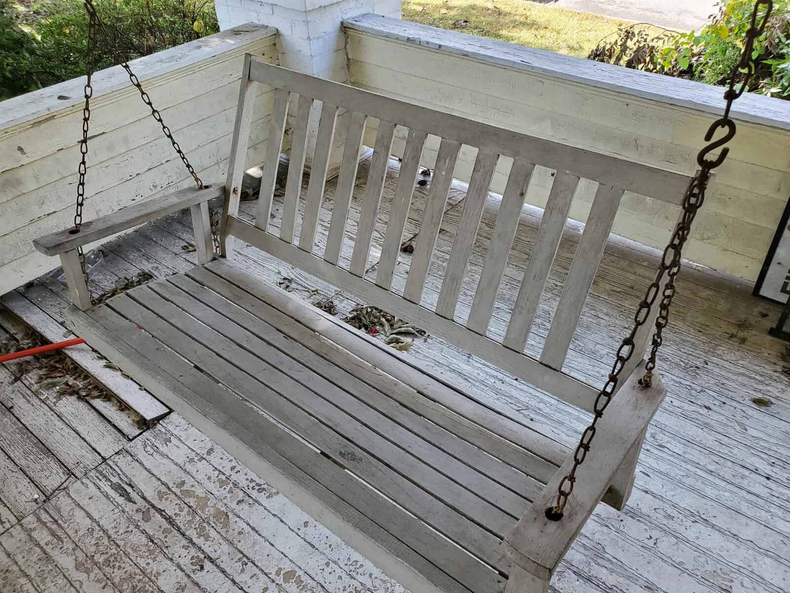 A wooden porch swing with visible wear and some leaves on it, attached to a white wooden porch by chains.