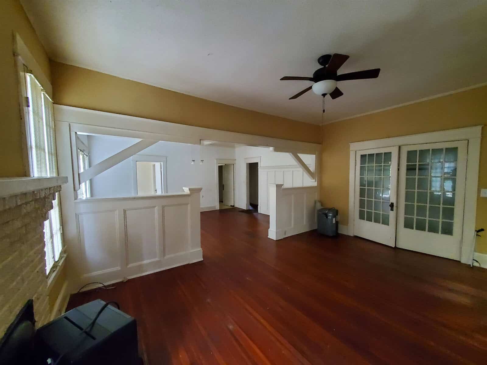 An empty living room with hardwood floors, beige walls, white wainscoting, a ceiling fan, and glass doors leading to another room.
