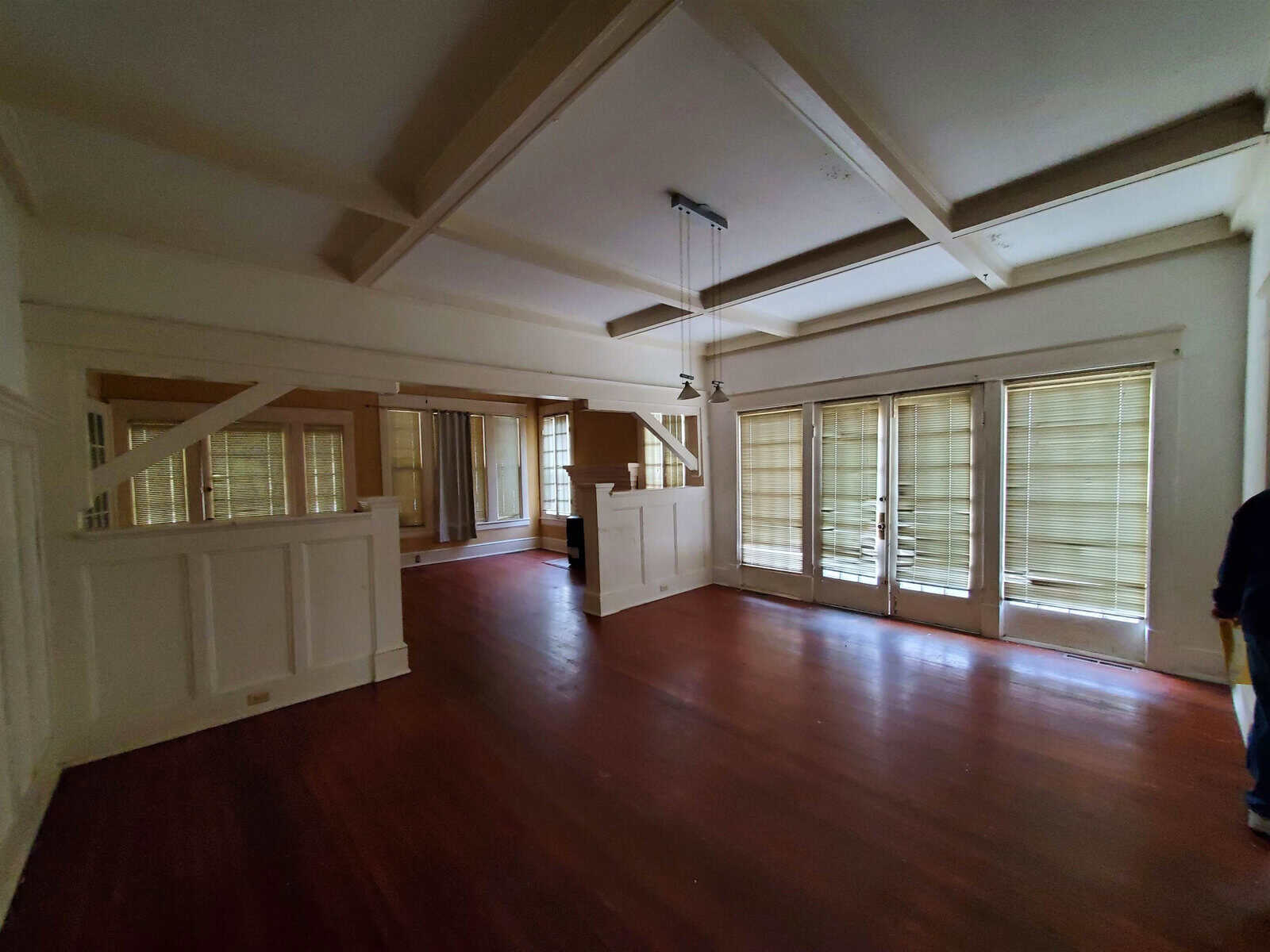An empty room with polished wooden floors, white walls, and multiple windows with closed blinds. the ceiling has wooden beams and there is partial view of a person on the right edge of the frame.