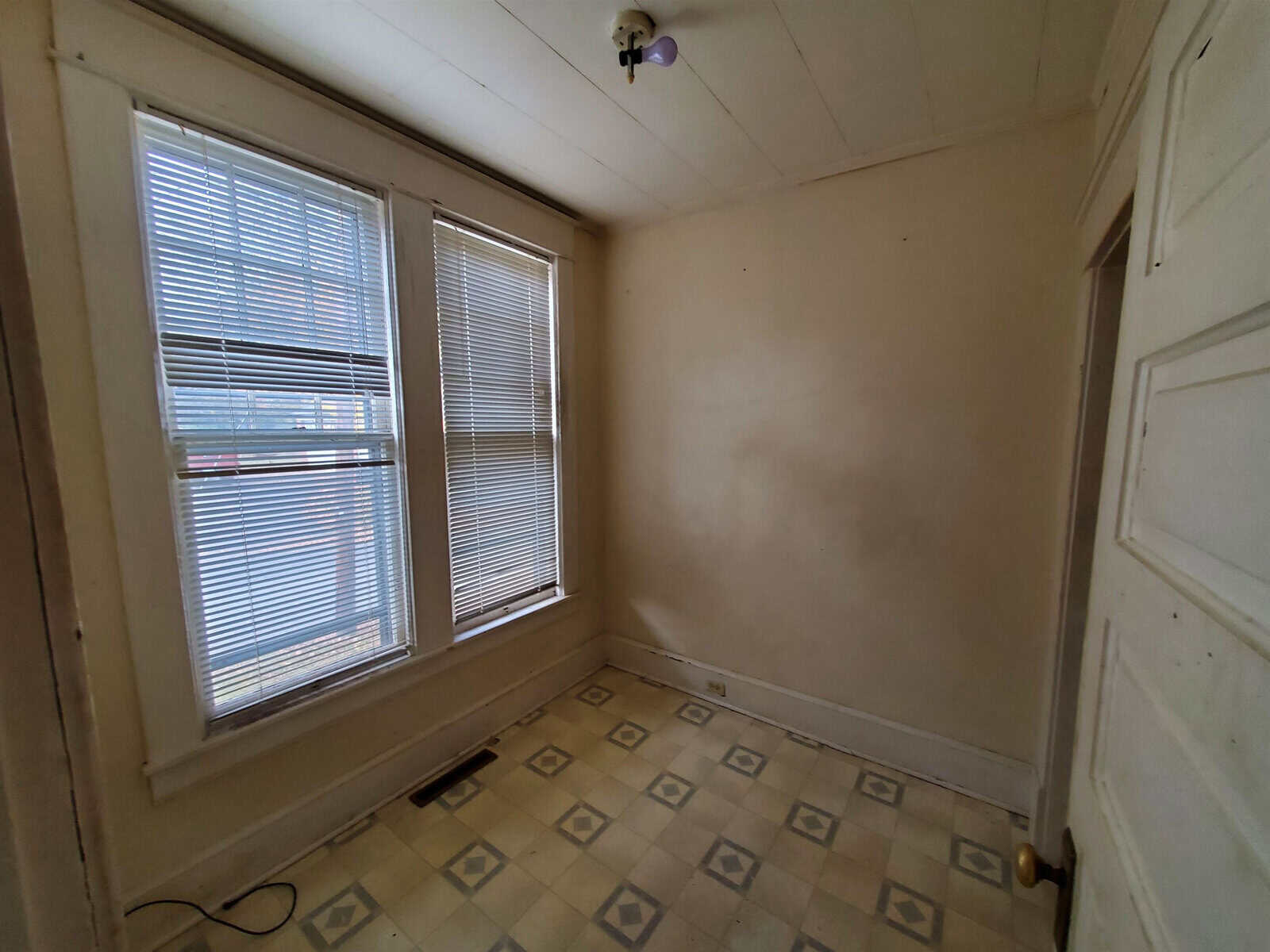 Empty room with worn white walls, a window with white blinds, and a tiled floor with a diamond pattern.