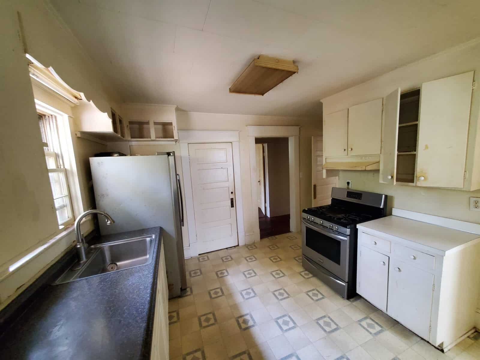A dated kitchen interior with white cabinetry, a freestanding gas range, a refrigerator, and a sink, showing signs of wear and in need of maintenance or renovation.