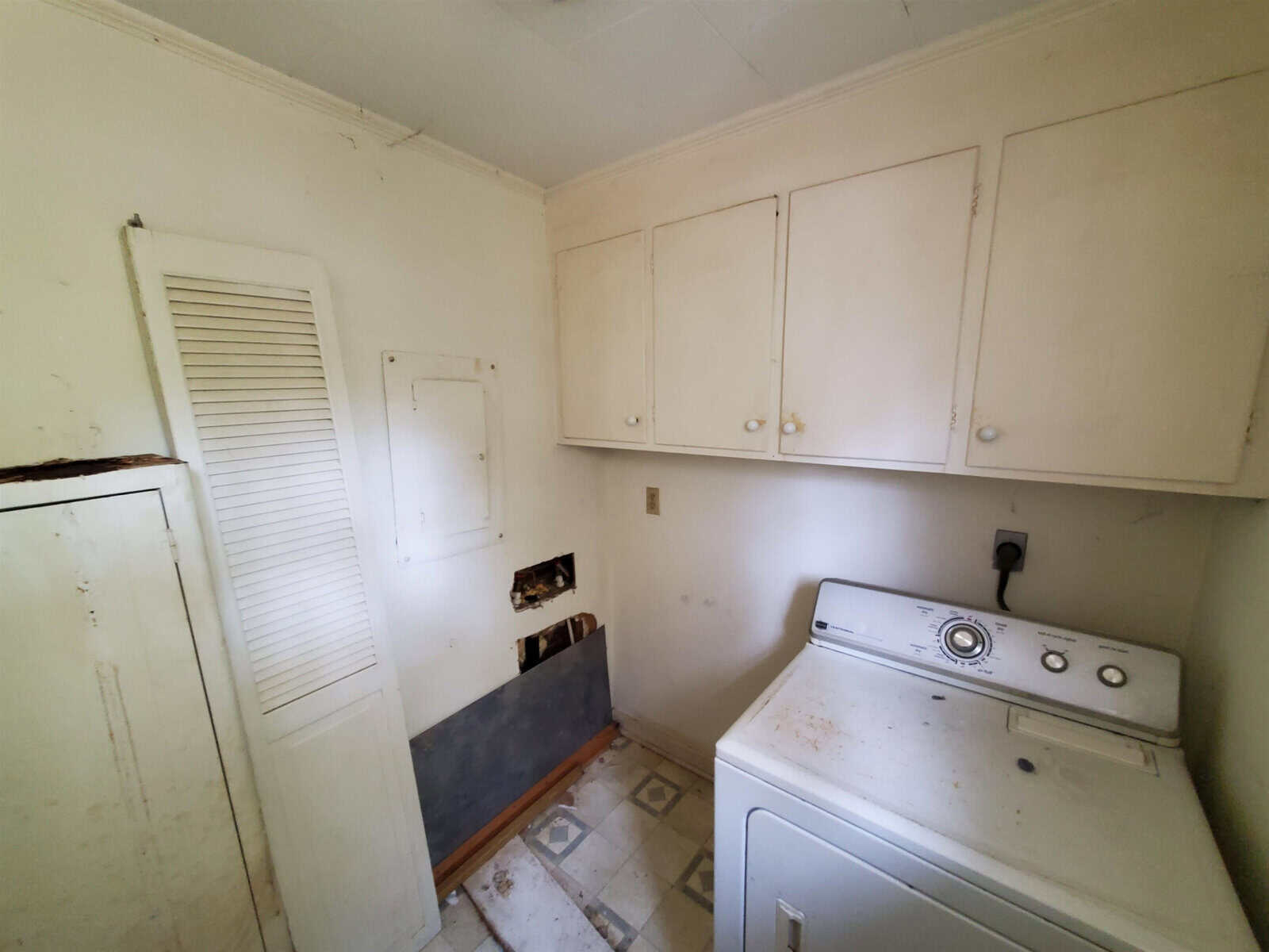 A neglected laundry room with a white washing machine, wall-mounted cabinets, and visible signs of wear and damage.