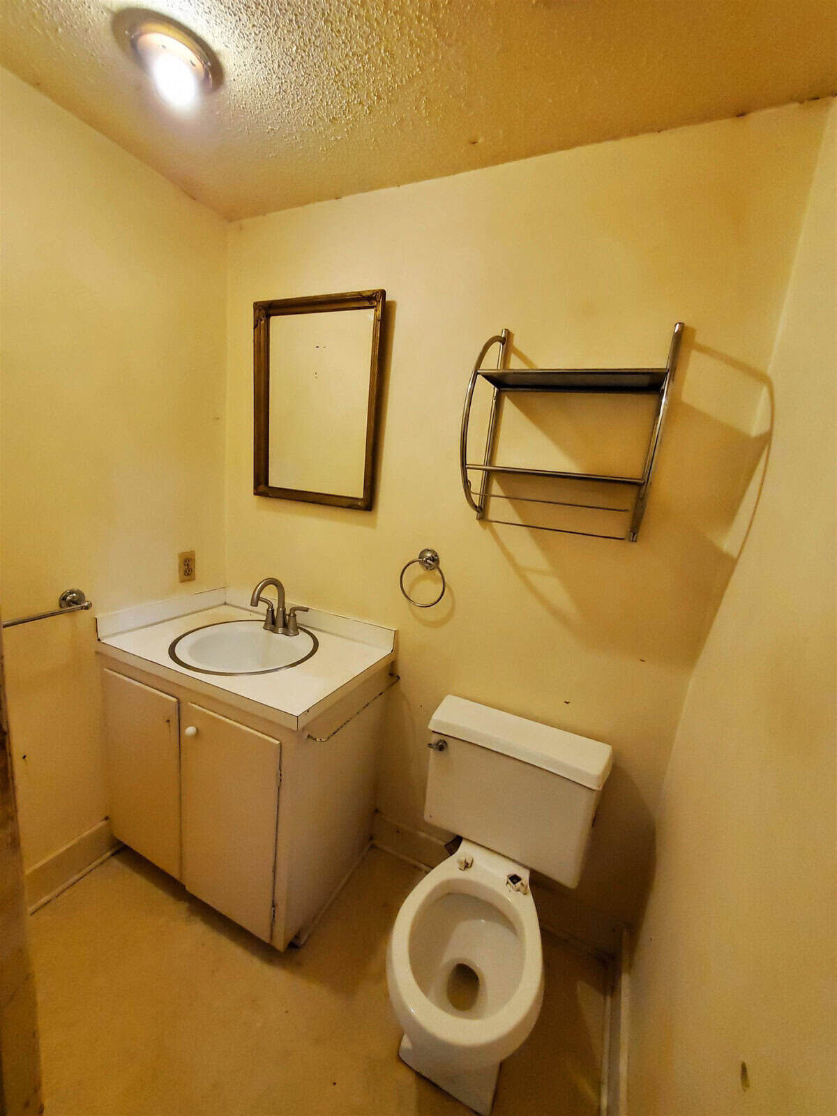 A small, narrow bathroom featuring a sink, toilet, and towel rack, with visible signs of wear and some discoloration on walls.