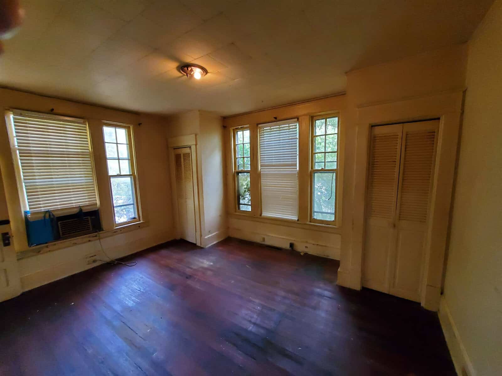 An empty room with wooden floors, two windows with blinds, a wall-mounted air conditioning unit, and a single lit ceiling light fixture.