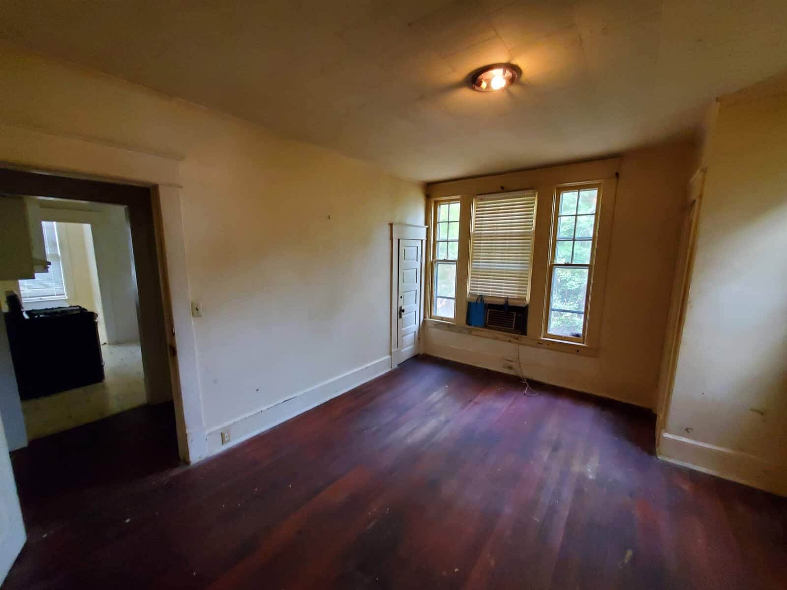 An empty room with hardwood floors, a single window with blinds, and a doorway leading to another room. the lighting is dim with one overhead fixture, and the walls appear in need of repainting.