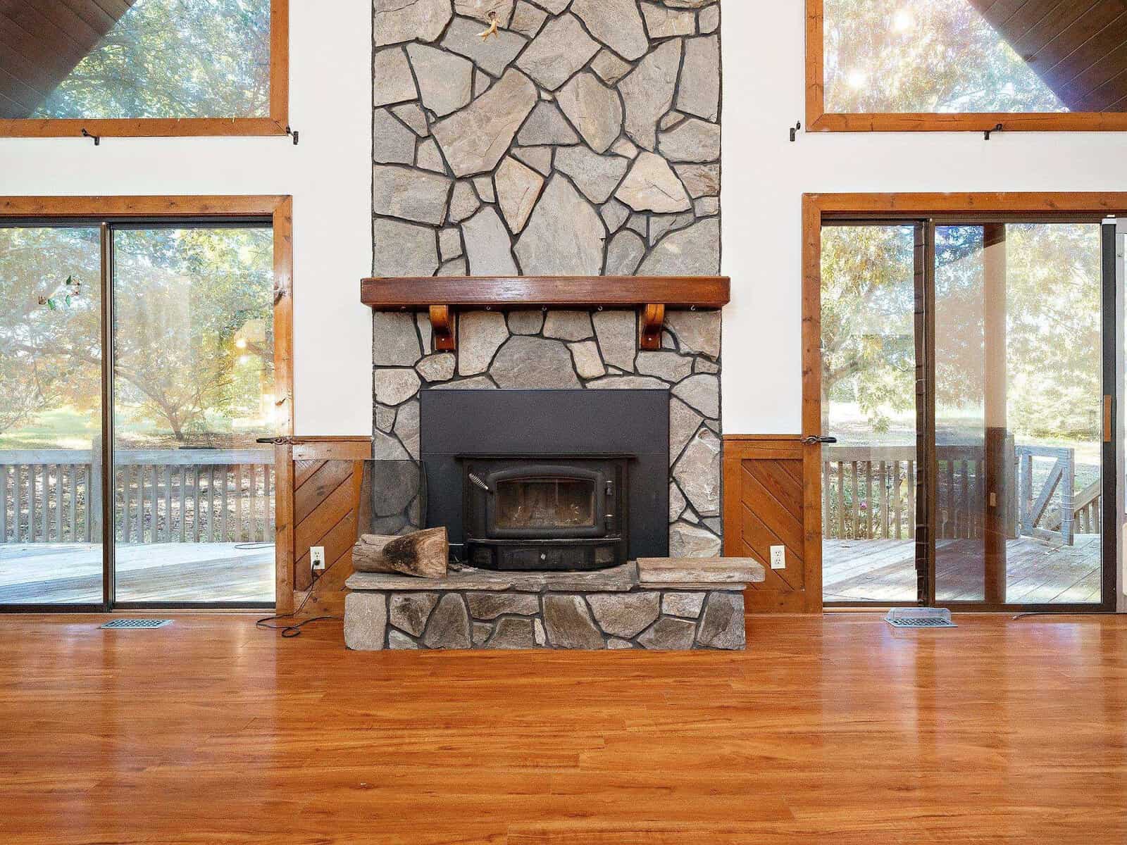 A cozy living room with a stone fireplace and a wood-burning stove. Large windows on either side of the fireplace reveal a spacious wooden deck and lush greenery outside. The room features a hardwood floor, and wooden beams frame the windows and ceiling.