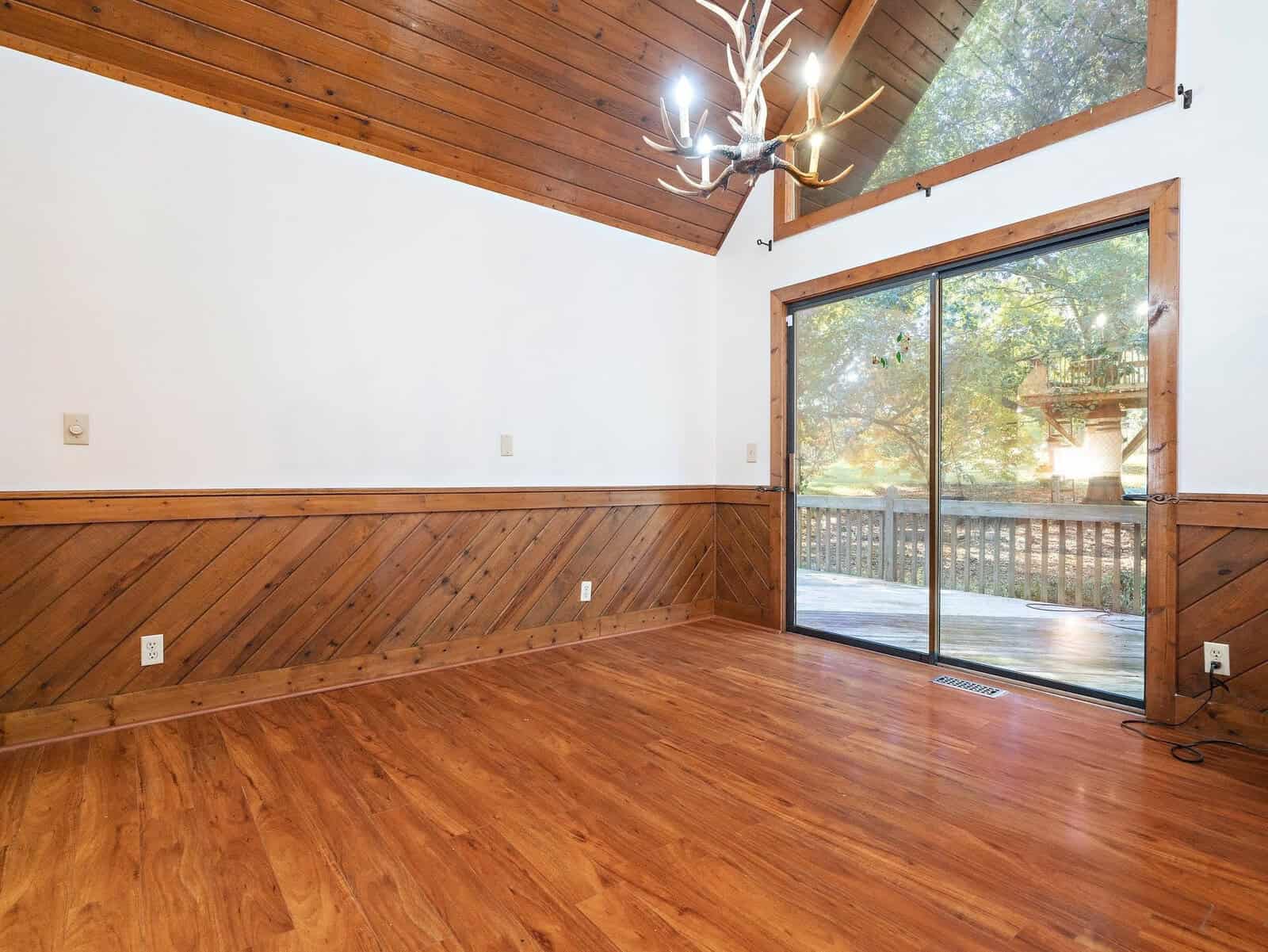A spacious room with wooden floors, paneled wooden walls, and a high angled ceiling. A large glass sliding door opens to a deck, offering a view of greenery outside. A modern chandelier with a rustic design hangs from the ceiling.