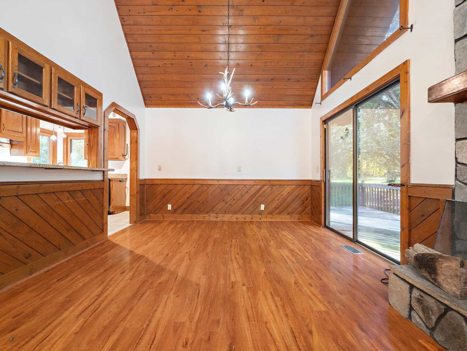 A spacious, empty room with wooden floors and a high, sloped wooden ceiling. A modern chandelier hangs in the center. There's a sliding glass door leading to a deck, and the walls have a mix of wood paneling and white paint. A stone fireplace is partially visible.