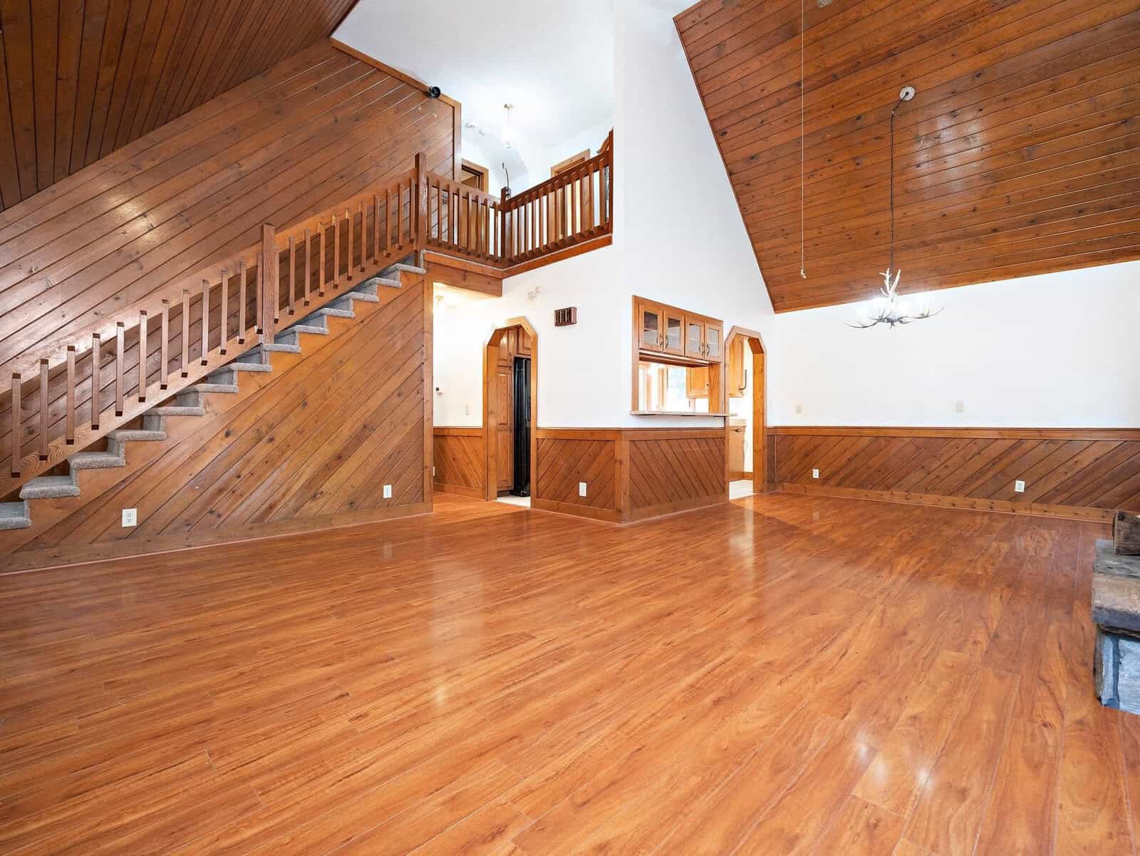 A spacious interior features wooden floors and paneled walls with a high, slanted ceiling. A wooden staircase ascends to an upper level, while wooden accents integrate throughout, framing entryways and railings. A chandelier hangs from the ceiling, adding a touch of elegance.