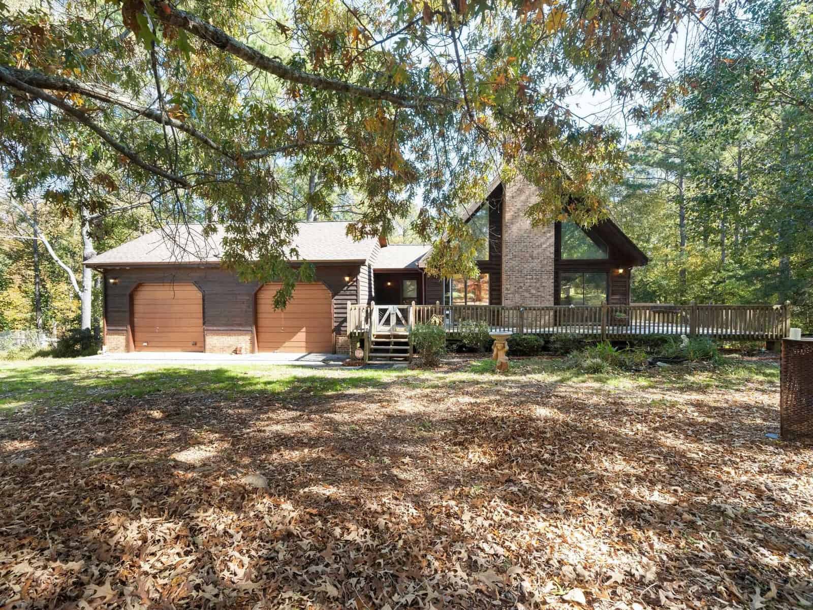 A rustic wooden house with a large wraparound deck and tall windows is surrounded by trees with autumn leaves on the ground. The house has a two-car garage on the left and a triangular windowed section on the right, blending seamlessly into the natural setting.
