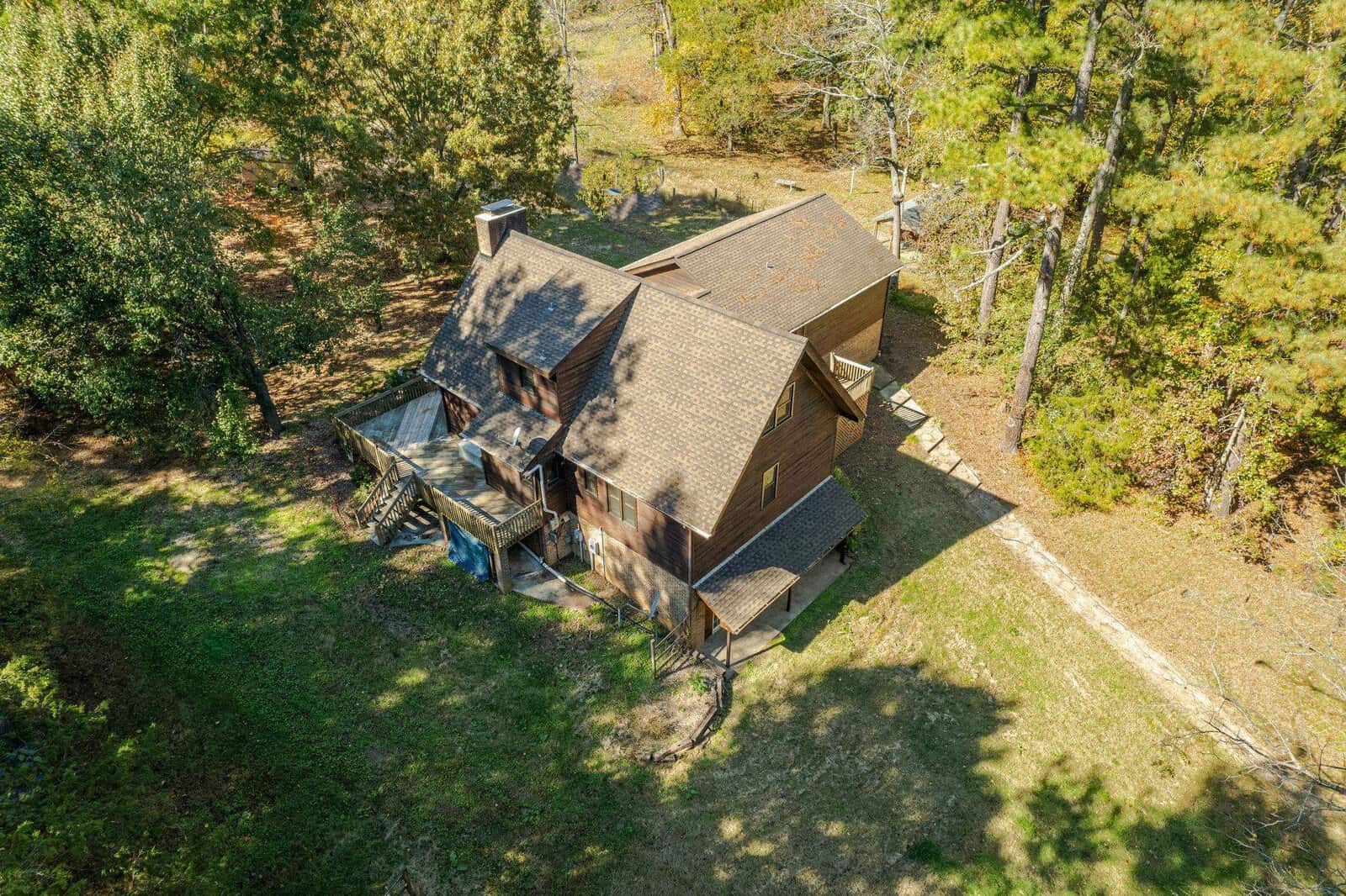 Aerial view of a two-story wooden house surrounded by trees and greenery. The house has multiple roof sections, a chimney, and a raised wooden deck with stairs leading to the ground. Shadows from the trees are cast around the property.