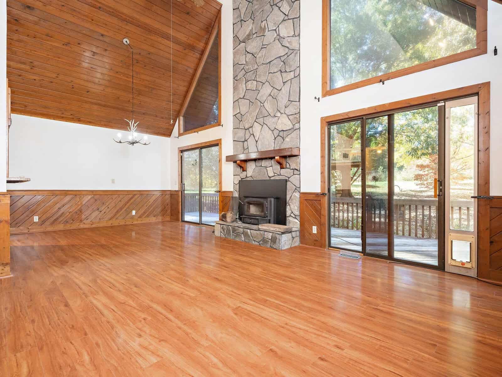 A spacious, empty living room with a high, vaulted wood ceiling, large windows, and a stone fireplace. The room features wooden flooring and sliding glass doors leading to an outdoor deck. Natural light floods the space, highlighting the serene, wooded surroundings.