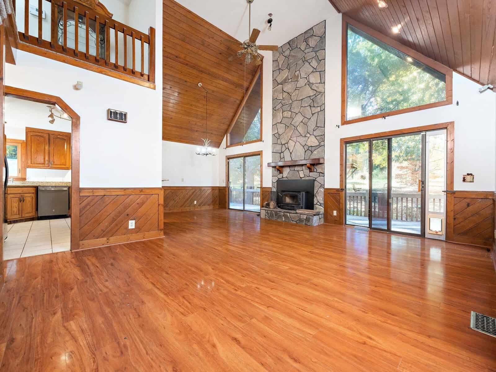 A spacious living room with high vaulted ceilings and large windows. The room features a stone fireplace, wooden flooring, and a wood-paneled ceiling. It opens to a deck through sliding glass doors. The kitchen is visible through an open doorway.