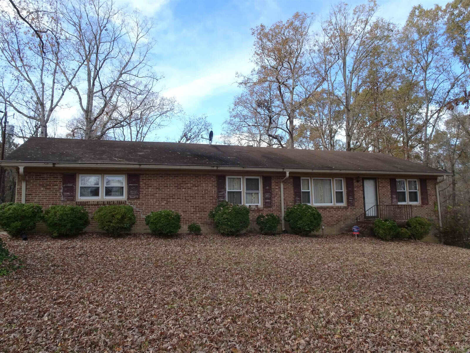 A single-story brick house with a dark roof surrounded by leaf-strewn lawn and bare trees, indicating autumn or winter season.