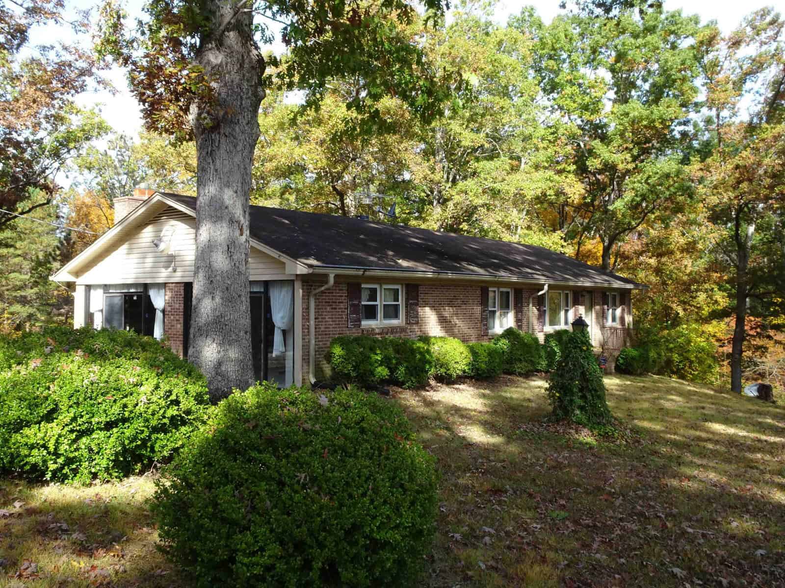 A single-story brick house surrounded by well-trimmed bushes and trees, with a front yard covered in fallen leaves.
