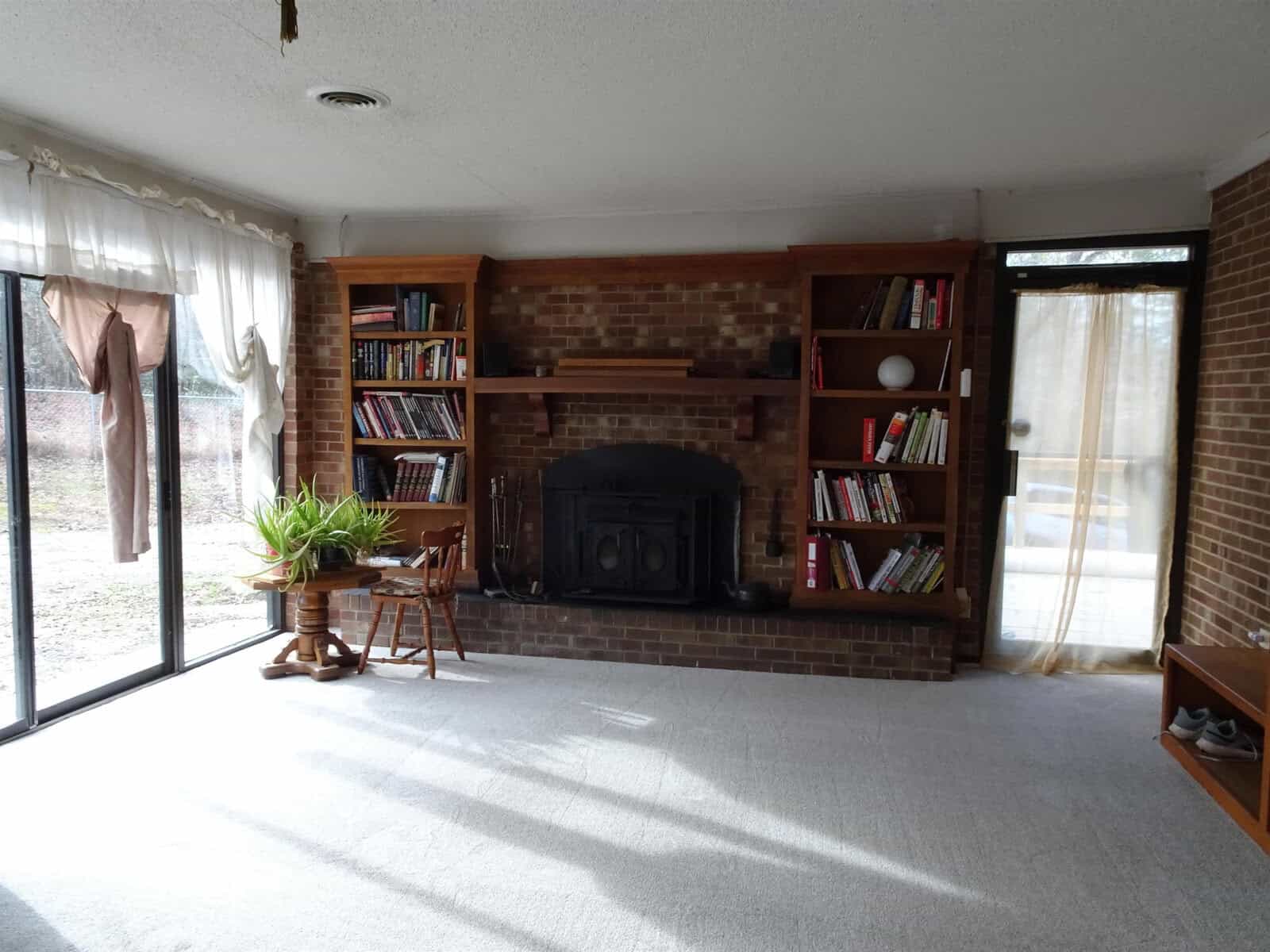 A spacious living room with a brick fireplace, bookshelves filled with books, a wooden rocking chair, a large window with curtains, and a glass door leading outside.