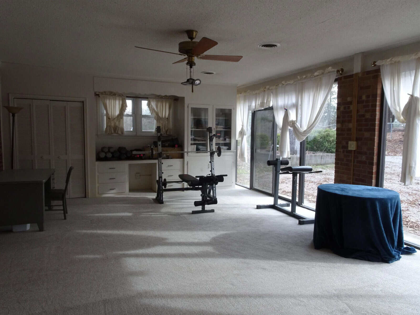 A home gym space with exercise equipment, including weights and benches, set up in a carpeted room with a ceiling fan and large windows draped with curtains.