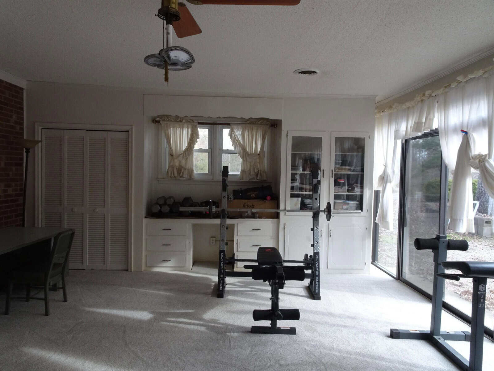 A home interior featuring an exercise area with weights and a bench, adjacent to a living space with white cabinetry and windows draped with sheer curtains.