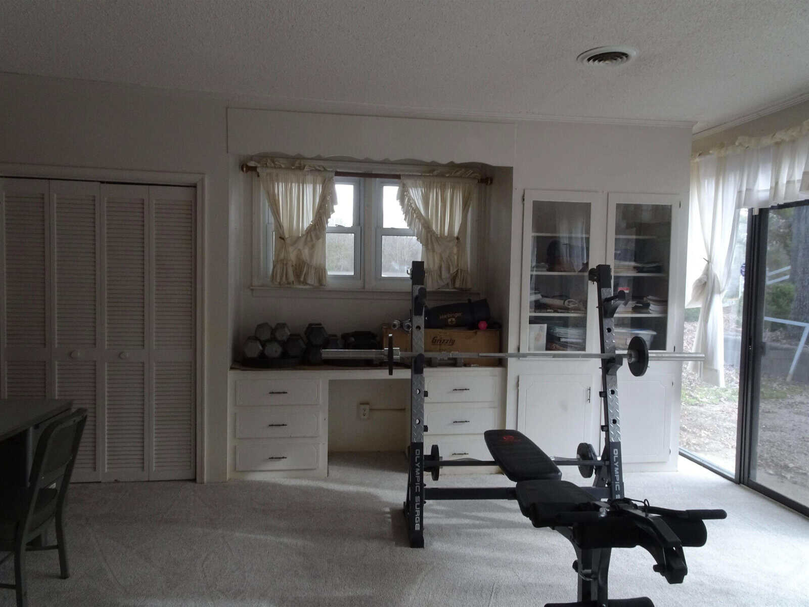 A home gym setup in a living space with exercise equipment including free weights and a bench, situated near a window with sheer curtains and built-in storage units.