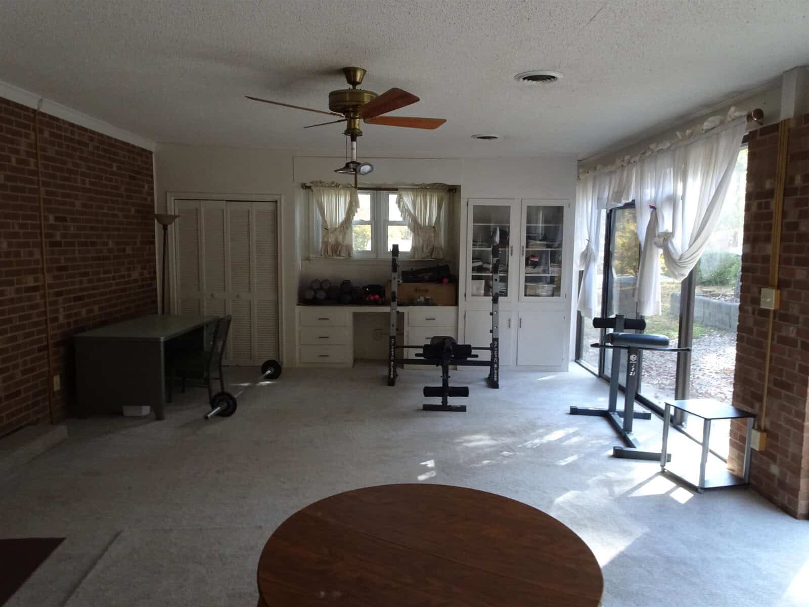 A spacious room with exercise equipment, including a weight bench and free weights, near a window with sheer curtains. the room features a ceiling fan, brick walls, white cabinetry, and carpeted flooring.