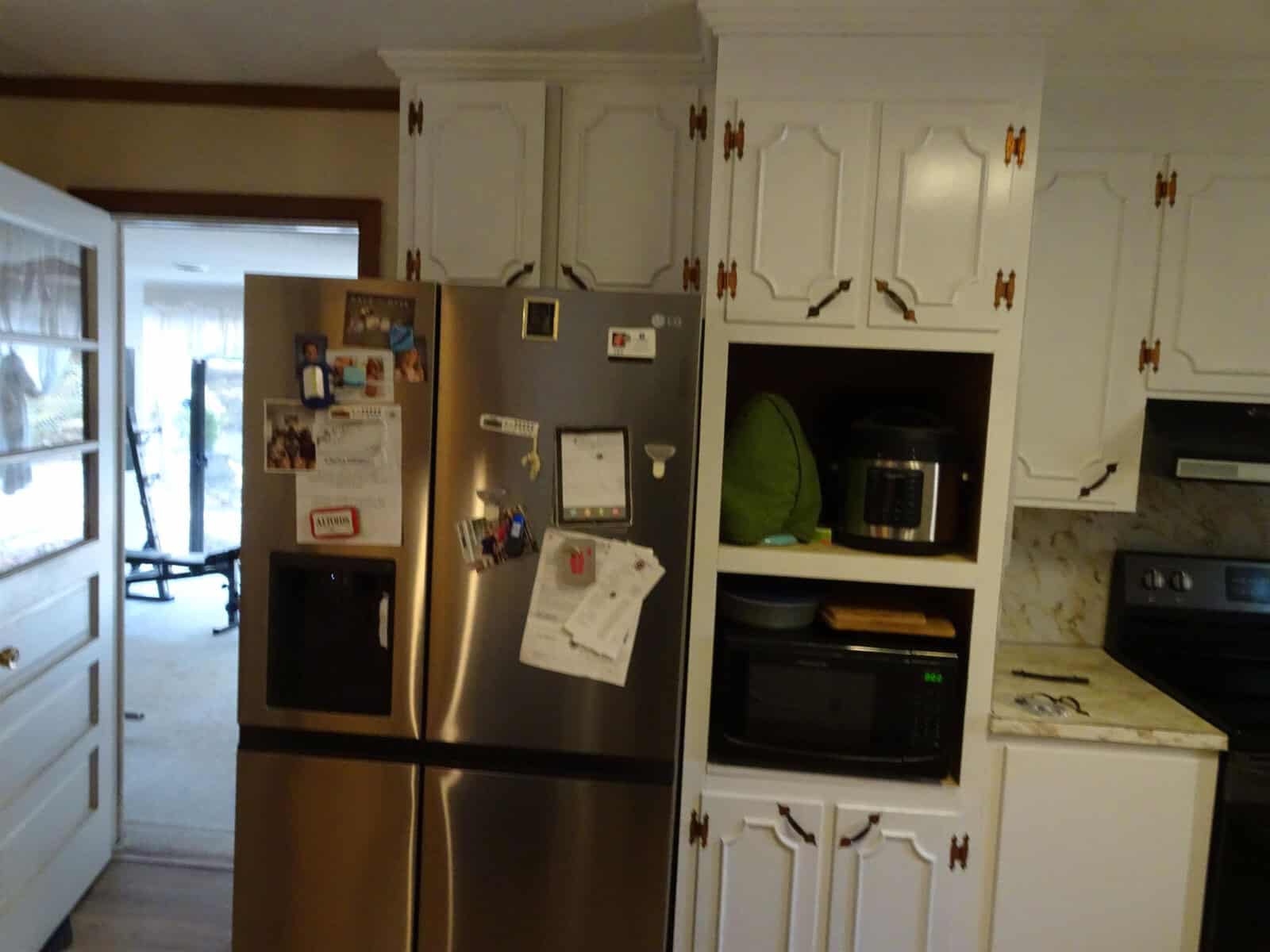 A kitchen interior showing a stainless steel refrigerator with magnets and papers attached to the door, flanked by white cabinets with black handles, and a glimpse of a stove and countertop appliances.