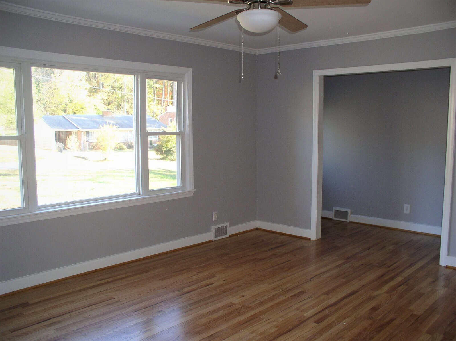 An empty room with hardwood floors, gray walls, white trim, and a ceiling fan, with sunlight streaming in through the windows.