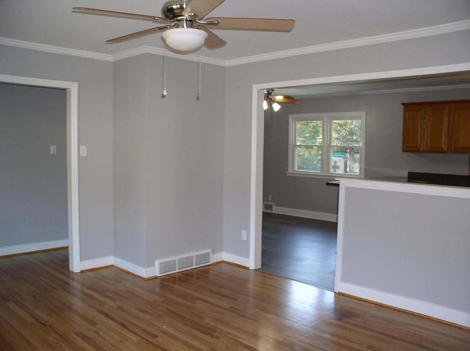 A clean and unfurnished room featuring hardwood floors, light gray walls, white trim, and a ceiling fan, with a view into an adjoining kitchen area.