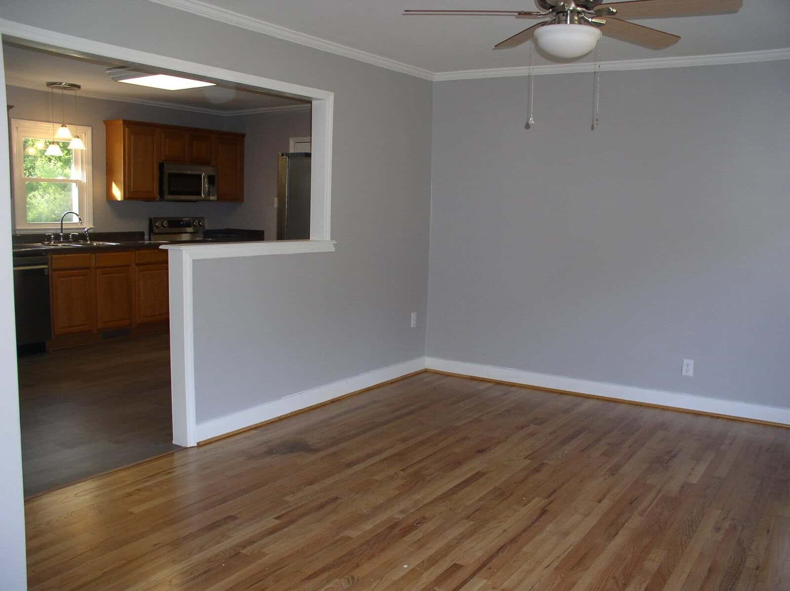 An empty room with wooden flooring, a half-wall partition, and a view into the adjoining kitchen area with wooden cabinets. a ceiling fan is mounted above.