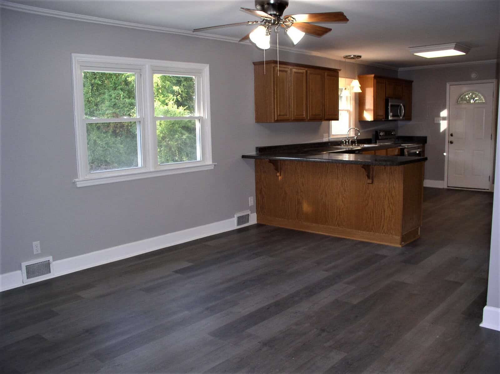 An empty room featuring a hardwood floor, with a kitchen area including wooden cabinets and black countertops, and a ceiling fan above. there are two windows providing natural light.