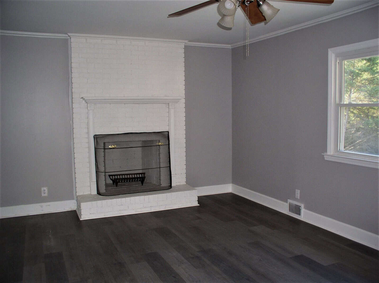 An empty room with gray walls and flooring featuring a white-painted brick fireplace and a ceiling fan.