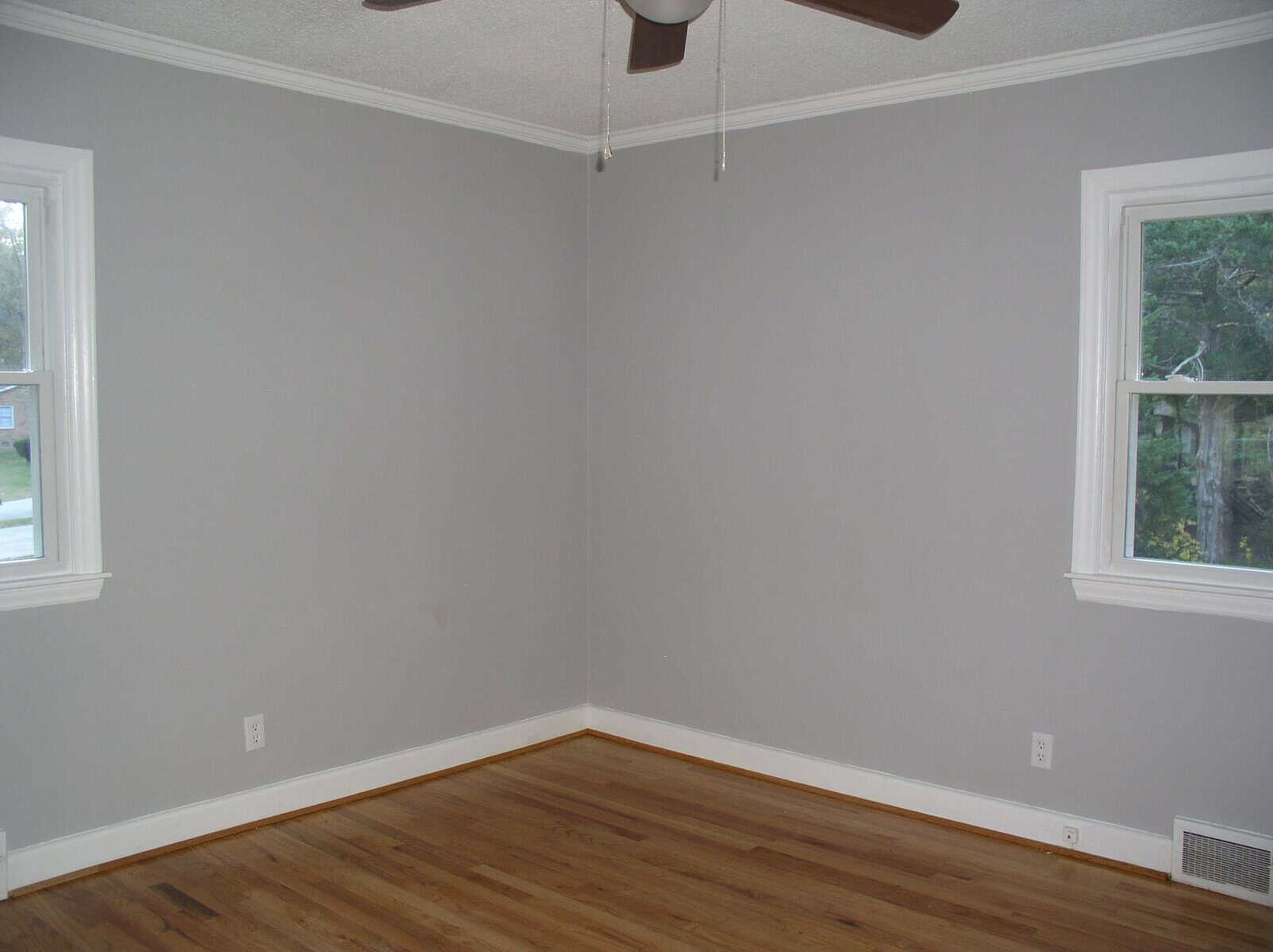 An empty room with grey walls, hardwood flooring, and a ceiling fan. two windows provide a view of the exterior greenery.