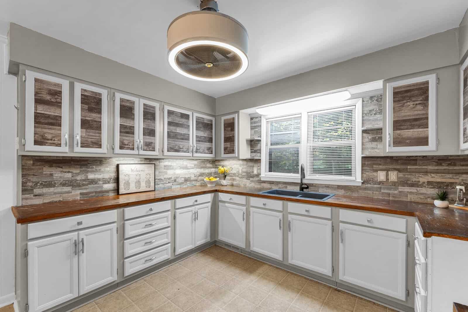 A well-lit, modern kitchen with white cabinetry, stone backsplash, and stainless steel sink beneath a window with horizontal blinds.