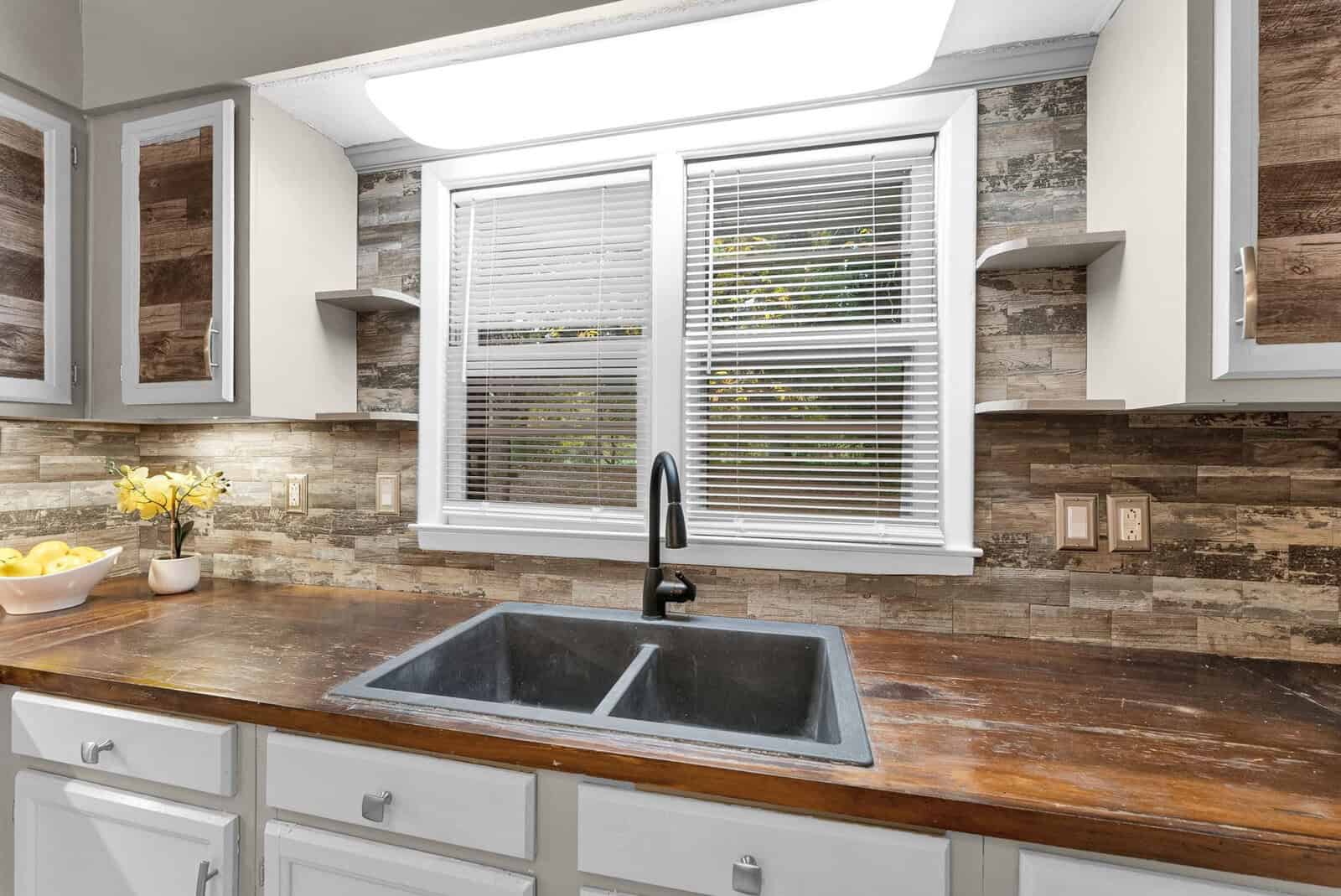 A modern kitchen interior featuring white cabinets, a stainless steel faucet with a black sink, wooden countertops, and a stone tile backsplash, with natural light coming in through a window with white blinds.