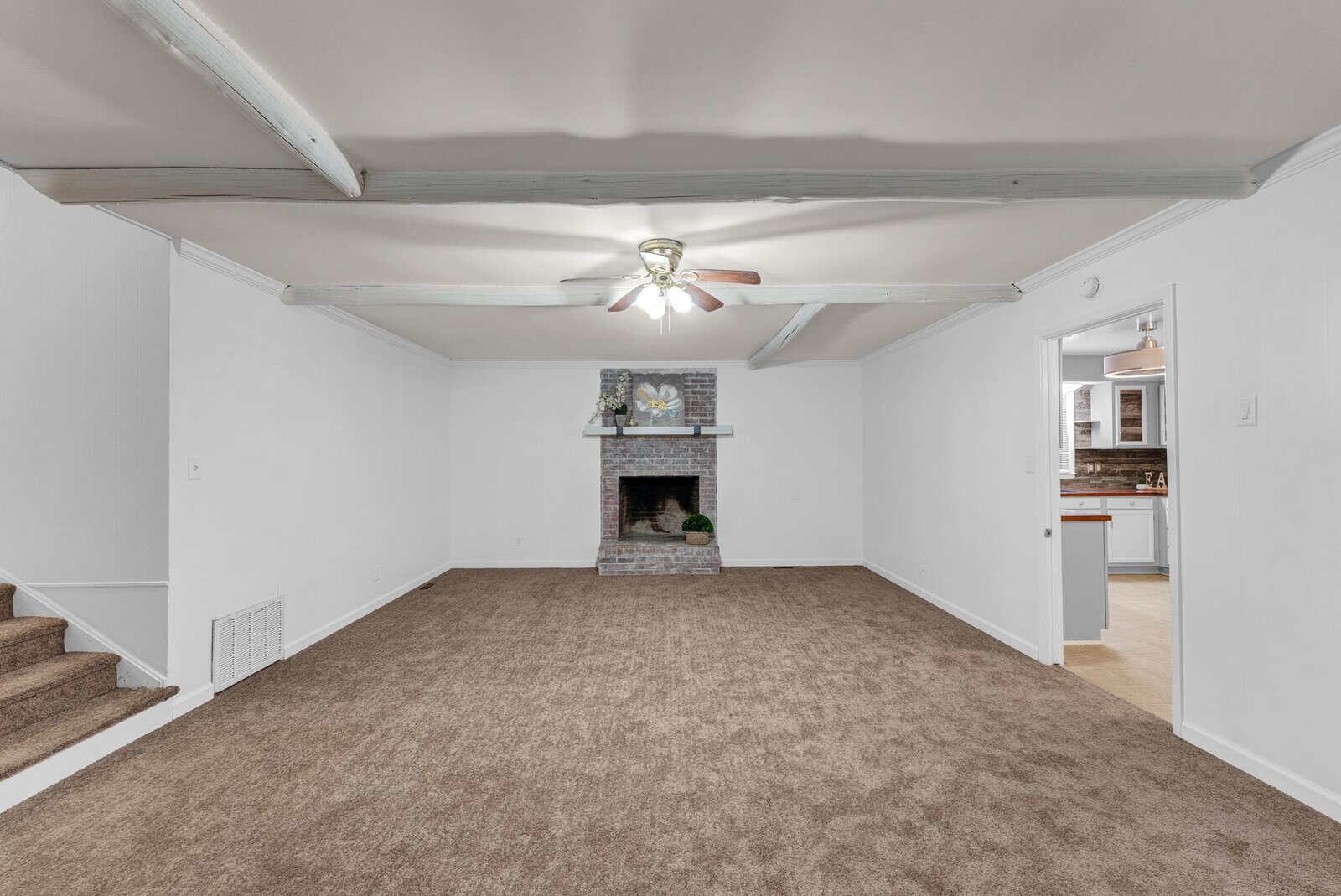 A spacious, empty room featuring a beige carpet, white walls, a fireplace with a decorative mantel, a ceiling fan, and a partial view into an adjoining kitchen.