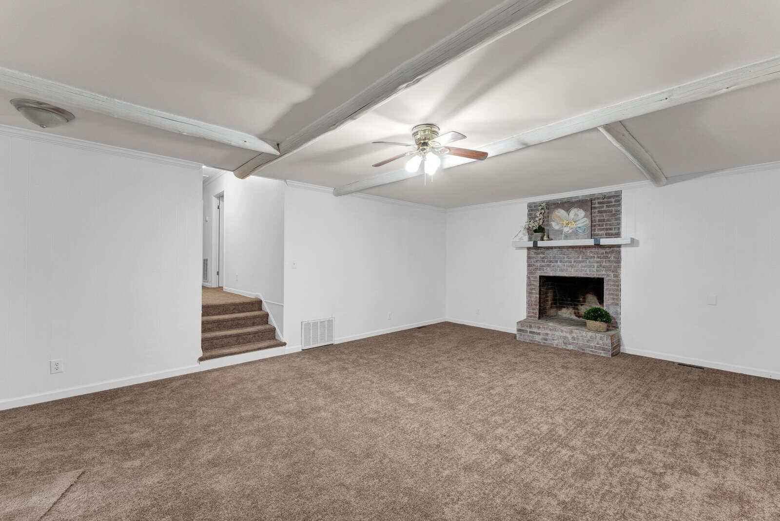 A spacious, empty room with white walls and a brown carpet featuring a brick fireplace and a ceiling fan, with a staircase leading to another level.